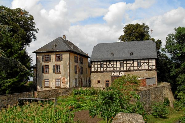 Blick auf das Schloss mit schlichtem Wohntrakt in L-Form und einem Wirtschaftsbebäude mit Fachwerkaufbau. Davor wird ein Nutzgarten von einer Steinmauer gerahmt. Rund um das Areal verläuft ein Weg vorbei an Bäumen.