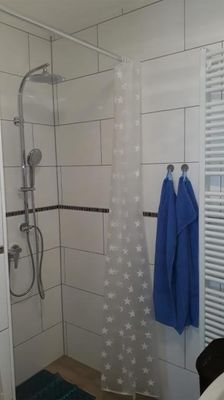Im hell gefliesten Badezimmer befindet sich eine Dusche mit Duschvorhang. Daneben hängen zwei blaue Handtücher und ein Handtuchheizkörper.