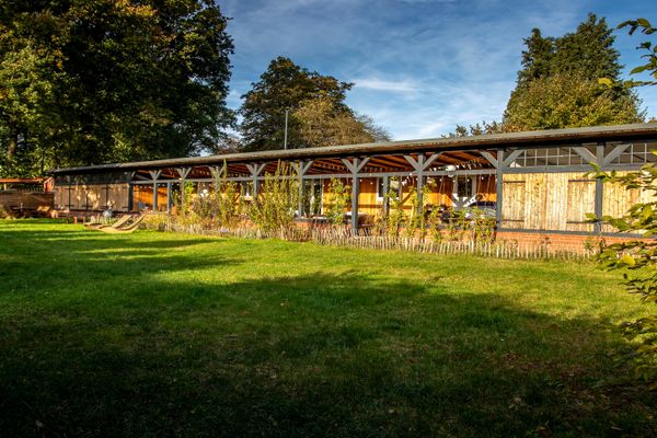 Außenaufnahme eines langgestreckten Gebäudes mit heller Holzfassade und einer langen, überdachten Veranda, die von einer dunklen Holzkonstruktion getragen wird. Davor erstreckt sich eine weitläufige, von der Sonne beschienene grüne Rasenfläche, die durch einen niedrigen, rustikalen Holzzaun von der Terrasse abgetrennt ist.
