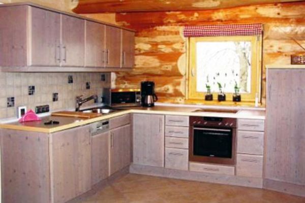 Eine L-förmige Holz-Küchenzeile mit Mikrowelle steht vor einer Wand aus Naturstammholz. Am Fenster ein hochgezogenes, rotkariertes Innenrollo.