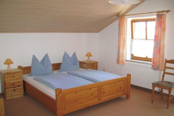 Doppelbett und  zwei Nachttische in Kiefer. Leichte Mansarde, helle Vorhänge vor dem Fenster. Neben dem Fenster steht ein Stuhl.