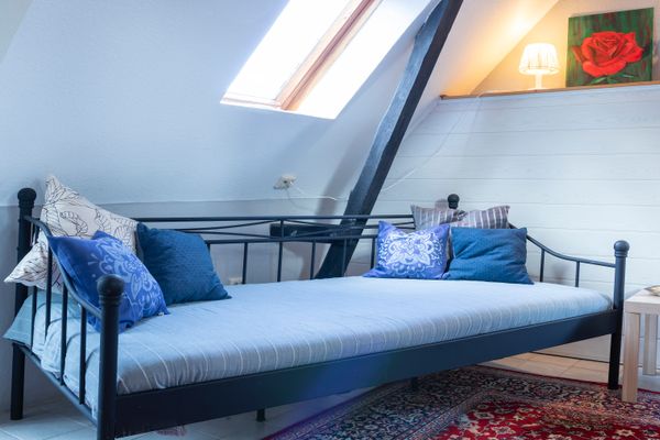 Ein Raum mit einer weißen Dachschräge und dunklen Holzbalken. Im Fokus steht ein schwarzes Metallbett mit einer hellblauen, gestreiften Matratze und mehreren blauen sowie grauen Kissen. Im Hintergrund ist ein Heizkörper an der Wand zu sehen, während durch ein kleines Dachfenster oben rechts Tageslicht einfällt.