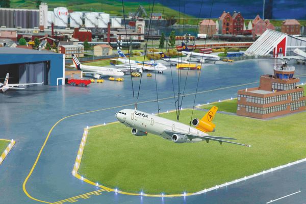 Mini-Flughafengelände mit Terminal und Hangar. Einige Flugzeuge befinden sich am Boden. Ein Flieger ist kurz nach dem Start in der Luft.