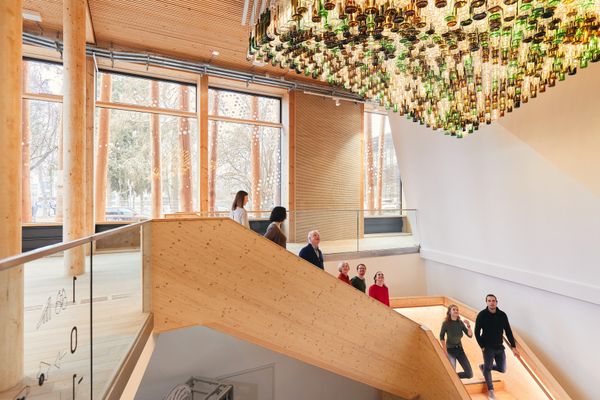 Über lichtdurchflutendem Treppenaufgang hängen unzählige Flaschen ordentlich aufgereiht von der Decke. Die daruntergehenden Menschen schauen staunend nach oben.