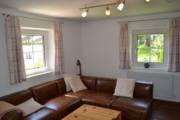 Wohnzimmer mit braunem Ecksofa vor zwei weißen Sprossenfenstern mit hellen, halbhohen Vorhängen; davor ein Tisch aus Kiefernholz, hinter dem Sofa eine Gitarre.