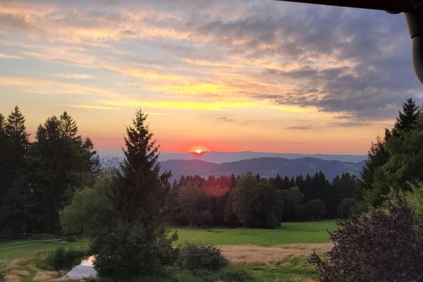 Ein traumhafter Sonnenuntergang beleuchtet eine wunderschöne Wald- und Hügellandschaft. Im Vordergrund erstreckt sich eine Wiese mit einem Teich, Bäumen und Sträuchern.