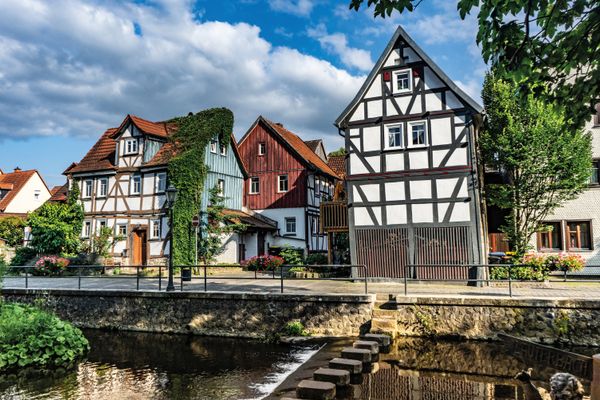 Fachwerkhäuser säumen das Ufer der Lauter, die durch das hessische Lauterbach im Vogelsberg fließt. Vor den Häusern verläuft eine schmale Straße, die an einem Uferkai mit Metallgeländer endet, denn hier verläuft etwas tiefer der Fluss Lauter. Die Kaimauer ist mit ein paar Stufen unterbrochen, die zu Schrittsteinen führt. Die Steine queren einmal den Fluss. Es ist herrliches Wetter.
