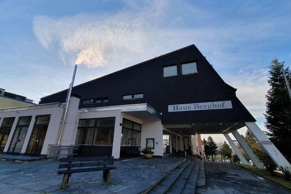 Drei schrägstehende, eckige Säulen tragen den überdachten Eingangsbereich einer Apartmentanlage, der mit dunklem Holz vertäfelt ist. Über dem Eingang prangt der Schriftzug „Haus Berghof“.