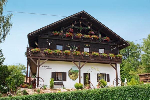 Ein Haus mit einem gemauerten Untergeschoss, dunklen Fensterläden, einer Wandmalerei und blumengeschmückten Balkonen. Ober- und Dachgeschoss sind mit dunklem Holz verkleidet und Geweihen dekoriert.