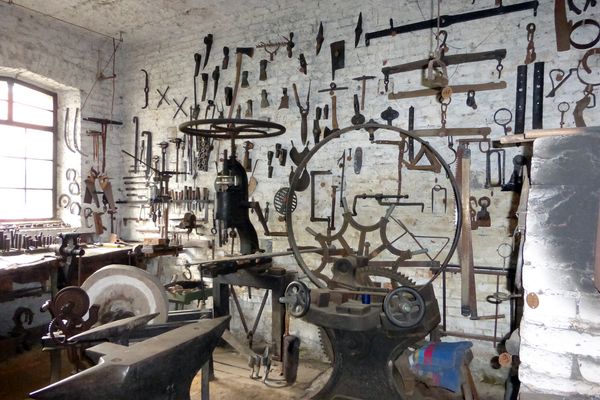 Blick in eine alte Schmiede. Viele Metallwerkzeuge hängen an der Wand. Davor stehen große, metalleren Geräte mit Stellrädern, ein Amboss und ein Schleifstein.