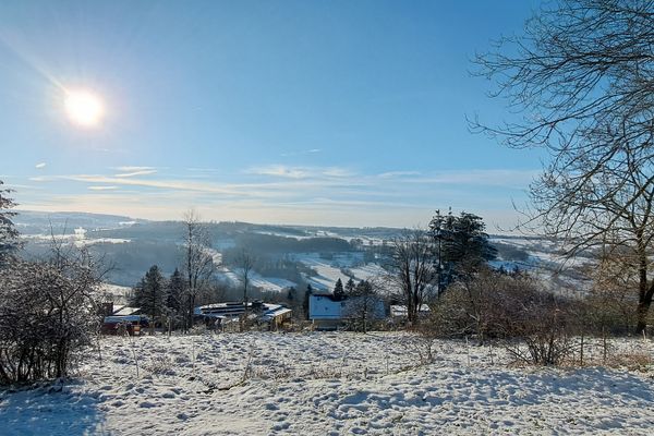 Der Blick geht hangabwärts über eine schneebedeckte Wiese auf eine winterliche Baumreihe und Häuser am Ortsrand. Dahinter tut sich ein weiter Blick über eine mit sanften Hügeln durchzogene Landschaft auf. Waldbereiche wechseln mit verschneiten Wiesen und Feldern. Das Licht der Sonne strahlt hell über der Landschaft und wirft lange Schatten.