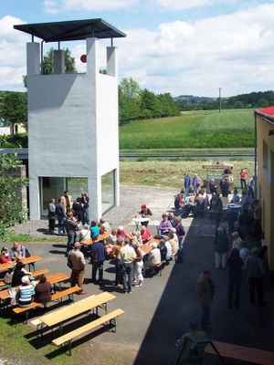 Rund um einen markanten, weißen Turm mit quadratischer Grundfläche und bodentiefen Fensternim Erdgeschoss stehen vielen Menschen. Manche sitzen auf Brauereigarnituren, es scheint eine gesellige Veranstaltung zu sein. Im Hintergrund ist hinter einer Straße ein Feld und angrenzend Bäume. In der Ferne ist weiter Mittelgebirgslandschaft zu sehen. Es ist schönes Wetter.