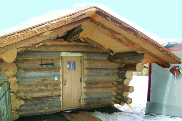 Holzblocksauna mit schneebedecktem Dach und großem Vordach. Schnee liegt auch neben dem Zugang und auf der dahinter liegenden Wiese. Neben der Blocksauna lehnt an einer Wand ein Schöpfeimer mit langem Stiel.