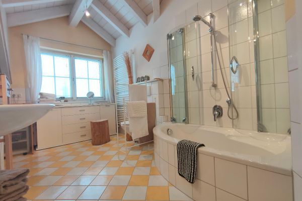 Ein Badezimmer mit orange-weißen Bodenfliesen unter einem offenen, hellen Dachstuhl. Es gibt ein Waschbecken, eine weiße Kommode unter einem Doppelsprossenfenster, einen Handtuchheizkörper, ein WC und eine Badewanne mit Glaswand.