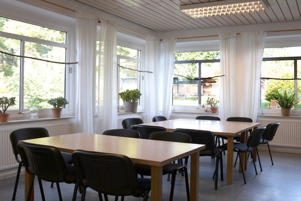 Ein heller Raum mit zwei rechteckigen Holztischen, um die mehrere schwarze Stühle gruppiert sind. An zwei Wänden befinden sich große Fenster mit weißen, transparenten Vorhängen und Topfpflanzen auf den Fensterbänken.