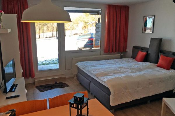 Ein graues Doppelbett mit roten Kissen steht gegenüber einer weißen Wohnwand mit TV. Am Fenster und vor der Terrassentüre hängen orangerote Vorhänge. Im Vordergrund ein Holzesstisch mit zwei Stühlen.