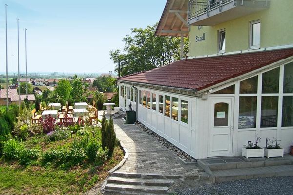Ein gepflasterter Weg trennt eine große, überdachte Terrasse von einem offenen Bereich mit vier Tischen, bedeckt mit Tischtüchern, zwischen Holzbänken. Von hier aus bietet sich ein weiter Blick über umliegende Häuser bis in die weite, ebene Landschaft.