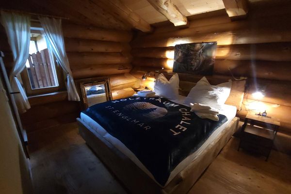 Schlafzimmer im Holzhaus mit großem Bett im massiven Holzrahmen, Wandbild und Fenster mit hellen Vorhängen. Auf der Tagesdecke steht: „Holztraum Chalet Sankt Englmar“.