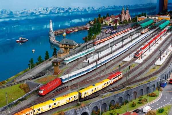 Modellbahnhof mit mehreren Zügen in verschiedenen Farben auf mehreren Gleisen. Hinter einem Gewässer grüßt eine verschneite Bergkette.
