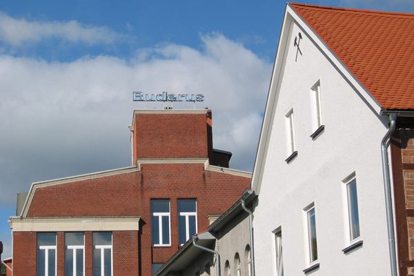 Der obere Ausschnitt eines Industriegebäudes aus rotem Backstein mit der blauen Leuchtreklame des Schriftzuges Buderus auf dem mittleren rechteckigen Gibelstück ist das Bildzentrum. Davor ist ein weiteres angeschnittenes Gebäude in weiß zu sehen, an dessen Stirnseite unter dem Dach zwei verschränkte Hammer als Symbol der Bergleute abgebildet sind.