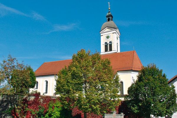 Hohe, bunte Laubbäume verdecken die Kirchenmauer, dahinter steht die Pfarrkirche Mariä Himmelfahrt in Wiesenfelden. Der markante Kirchturm ist mit Schindeln bedeckt.