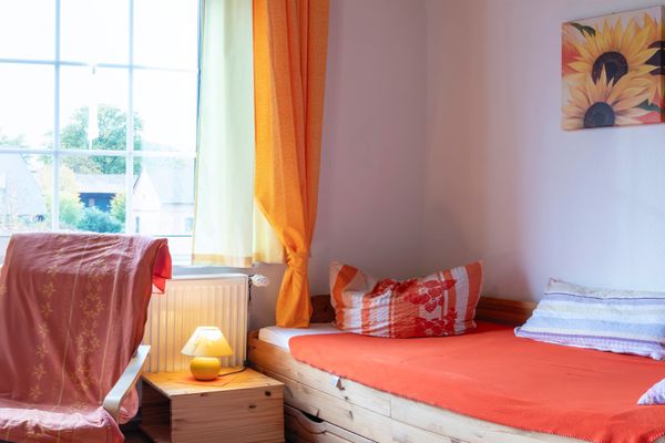 Einzelbett mit orangefarbener Bettwäsche, Fenster mit Vorhängen, Sessel und Nachttisch mit Lampe