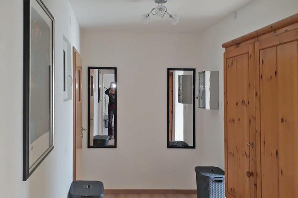 Im Flur stehen ein Holzschrank und zwei graue Kunststoff-Wäscheboxen. An der Wand hängen ein Bild sowie zwei lange, rechteckige Spiegel.