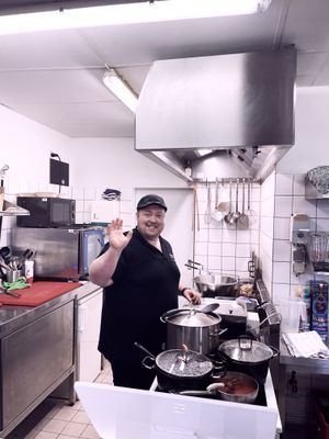 Auf dem Bild ist der Koch des Bistros in der Küche stehend zu sehen. Er winkt dem Fotografen zu.