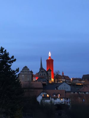 Die Silhouette der Burgenstadt Schlitz hebt sich deutlich vom blauen Nachthimmel ab. Im Zentrum leuchtet eine rote Kerze hell auf. Der Hinterturm wurde dazu mit einem roten Tuch verhüllt und hat eine beleuchtete Kerzenspitze erhalten. Ringsum sind historische Fachwerkbauten und steinerne Häuser unterschiedlichster Art zu sehen.