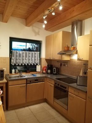 Unter einer Holzdecke mit freiliegenden Balken steht eine L-förmige Küchenzeile aus hellem Holz. Über der Spüle befindet sich ein Fenster mit Bistrovorhang.