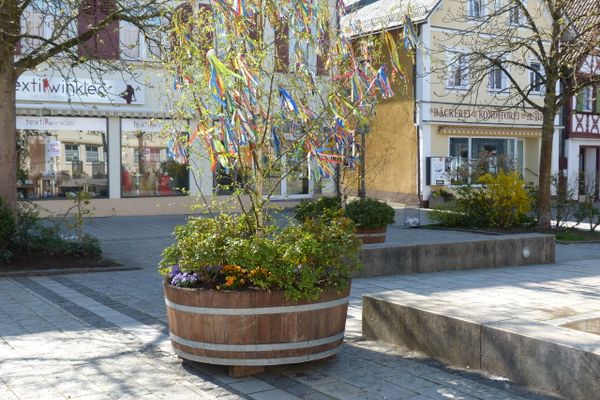 In einem großen Holztrog ist ein kleiner Baum, an dem viele bunte Bänder hängen.