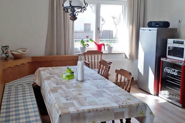 Im Raum stehen eine Holzeckbank, ein Tisch und drei Stühle. Gegenüber befinden sich ein hoher, grauer Kühlschrank und ein kleinerer, roter Getränkekühlschrank mit Glastür und der Aufschrift "Coca Cola".