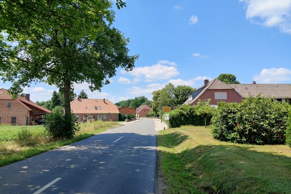 Straßenansicht am Ortseingang von Holxen mit asphaltierter Fahrbahn und seitlichem Grünstreifen. Links und rechts stehen Wohnhäuser mit roten Ziegeldächern sowie Gärten. Am rechten Straßenrand befindet sich ein gelbes Ortsschild, darüber ein blauer Himmel mit einzelnen Wolken.