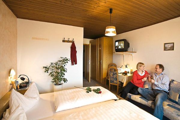 Auf dem Sofa gegenüber dem Doppelbett sitzt ein Pärchen und prostet sich mit Sekt zu. Daneben ein Tisch mit zwei Stühlen, darüber ein kleines TV-Gerät. Im Eingangsbereich ein Holzschrank hinter der Tür. Neben dem Bett eine Grünpflanze und drei Garderobenhaken an der Wand.