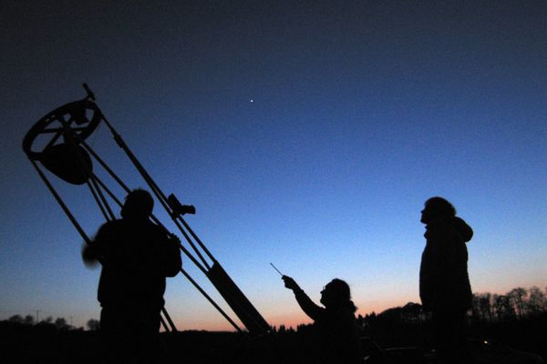 Der Himmel ist dunkel und klar, die Sterne leuchten hell. Im Vordergrund ist die Silhouette einer Frau und eines großen Teleskopes vor ein paar Bäumen zu sehen.