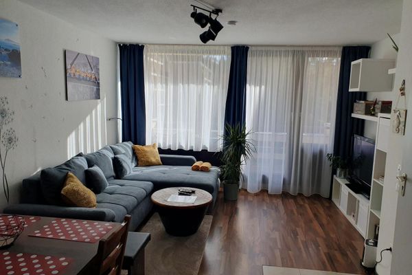 Vor einer Fensterfront mit weißen Gardinen und blauen Vorhängen stehen ein graues Ecksofa mit Kissen, ein dunkler runder Tisch mit Holzplatte und eine weiße Wohnwand mit TV. Im Vordergrund befinden sich ein dunkler Holztisch, eine Bank und ein Stuhl. Bilder und Dekomalerei zieren die Wand.