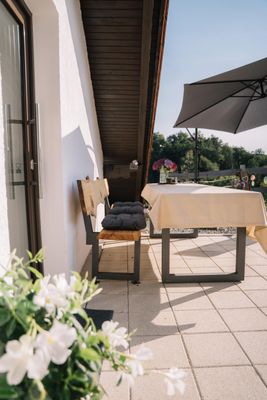 Auf der Terrasse mit hellen Fliesen steht eine Holzbank mit grauen Polstern versehen, davor ein großer Tisch mit heller Tischdecke. Ein großer Sonnenschirm steht vor der Sitzgruppe.