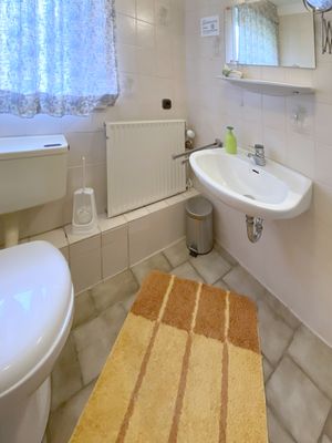 Bad mit Waschbecken, Spiegel, Fenster mit Vorhang, Heizkörper und WC sowie Teppich auf dem Boden