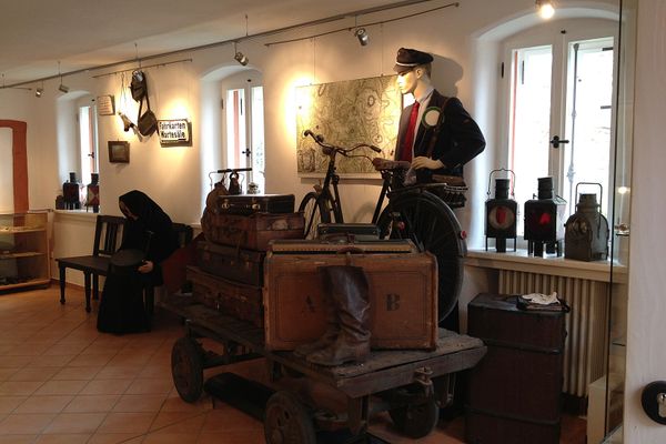 In einem weißen Raum mit braunen Fliesen geht der Blick auf ein Arrangement mit einem Zugwagen, der zwei auffällige rote Fahnen mit weißem Punkt an seiner Stirnseite hat und mit alten Koffern und einem alten Fahrrad bestückt ist. Dahinter steht eine Schaufensterfigur mit blauer Uniform. Rechts neben der Figur sind alte Rucksäcke über ein Geländer gehängt. An der Wand hängt ein Schild mit Aufschrift "Fahrkarten Wartesäle" und Pfeil nach links. Ein Signalhorn, eine Büchse und ein weiters Schild hängen an der Wand.