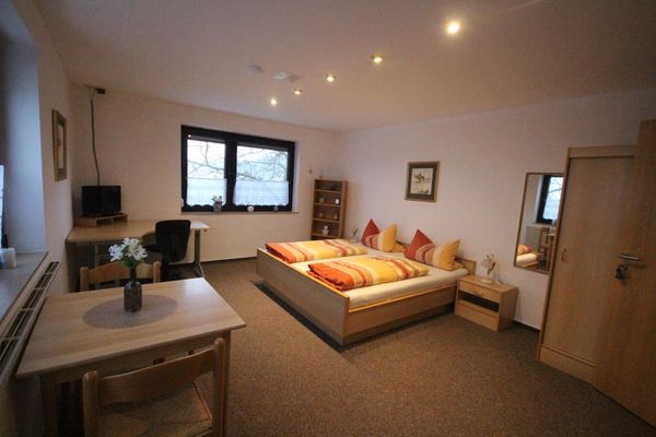 HilmerDoppelzimmer mit Holzmöbeln und Teppichboden. Schreibtisch mit TV, viereckiger Tisch und zwei Stühle vor dem Fenster. Spiegel, Eckregal, Bistrogardinen und zwei Wandbilder.