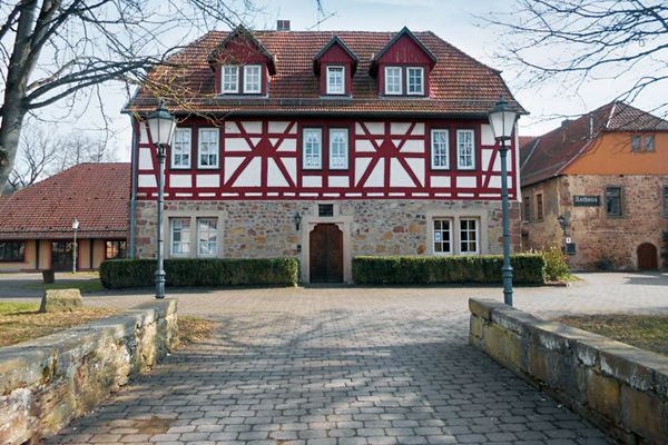 Ein prächtiger, roter Fachwerkbau, der auf einem steinernen Erdgeschoss sitzt, bildet das Zentrum des Bildes. Ein gepflasterter Weg, gesäumt von einer Mauer, führt direkt darauf zu. Rechter Hand ist ein weiterer, steinererner Bau mit orangem Giebel zu sehen. Das ist das Ratahaus. Linker Hand schließt sich ein neuerer, etwas niedrigerer Bau an.