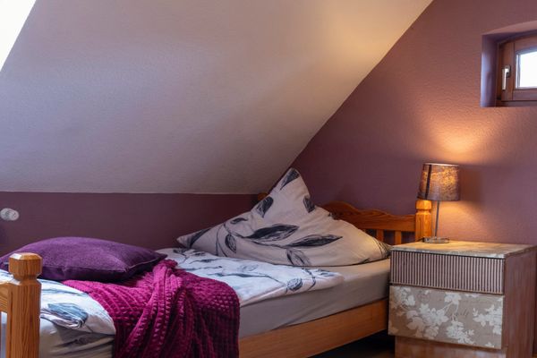 Ein Raum mit violetter Wand und weißer Dachschräge, durch die Tageslicht aus einem Dachfenster fällt. Im Mittelpunkt steht ein hölzernes Einzelbett mit blattgemusterter Bettwäsche, einem violetten Kissen und einer violetten Kuscheldecke. Rechts neben dem Bett befindet sich ein gemusterter Nachttisch mit einer eingeschalteten kleinen Lampe.