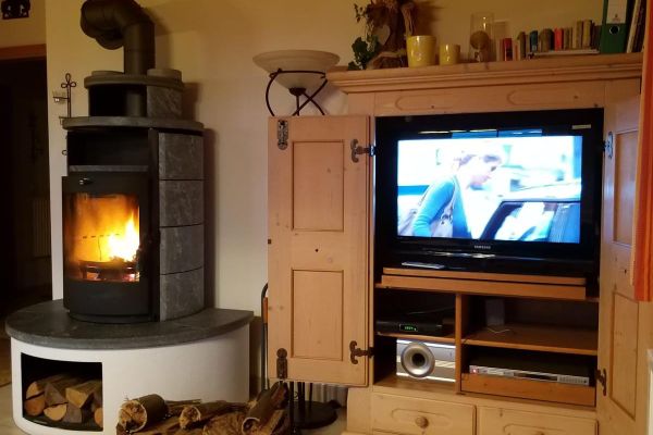 Fichtenholzschrank mit TV und Stereoanlage. Im daneben stehenden Kachel-Kaminofen mit Sockel brennt ein helles Feuer. Zwischen Schrank und Ofen befinden sich Kaminbesteck, eine Deckenleuchte und ein Korb mit Holz.