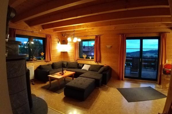 Unter einer Holzdecke mit freiliegenden Balken steht ein graues Ecksofa mit Zusatzhocker und Holztisch vor einem Kachel-Kaminofen. Die Wände sind ebenfalls aus Holz. Zwei Fenster und eine Doppelbalkontüre eröffnen einen herrlichen Blick in die Landschaft.