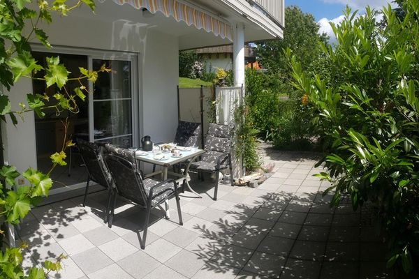 Unter einem Balkon liegt eine sonnige Terrasse, umgeben von hohen, üppigen Sträuchern. Auf der Terrasse stehen ein gedeckter viereckiger Frühstückstisch und vier Gartenstühle mit Sitzauflagen.