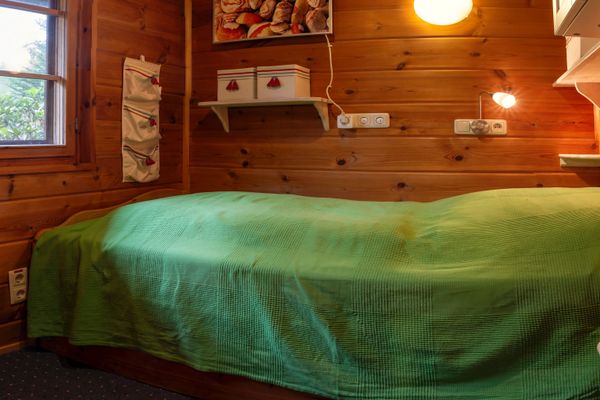 Einzelbett mit grüner Tagesdecke in einem holzverkleideten Raum, mit Fenster, Wandlampen und kleinen Regalen.