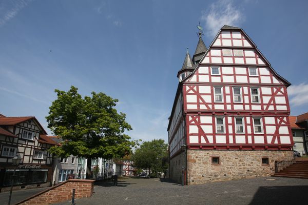 Rechter Hand erhebt sich ein großer roter Fachwerkbau mit geschiefertem Dach, zwei Spitzgauben und mittigem Turm mit Wetterfahne. Das Fachwerk-Rathaus wurde auf einem Sandsteinsockel gebaut. Vor dem Rathaus geht es hangabwäts über eine Treppe und eine Ebene mit Basaltpflastersteinen zu weiteren, tieferliegenden Fachwerkbauten.