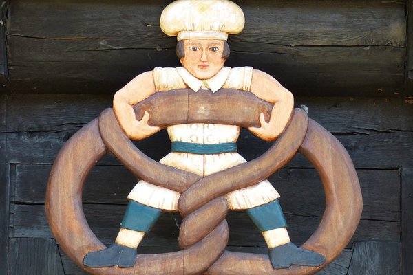 Auf dem Bild ist ein Schild zu sehen. Das Schild bildet einen Bäcker mit Kochmütze und Schürze ab, der sich an einer überdimensional großen Breze festhält und in dieser steht.