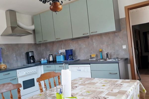 Im Raum steht eine L-förmige Küchenzeile mit hellblauen Schranktüren, ausgestattet mit mehreren Elektrogeräten. Daneben befinden sich ein Tisch mit gemusterter Tischdecke und zwei Holzstühle.