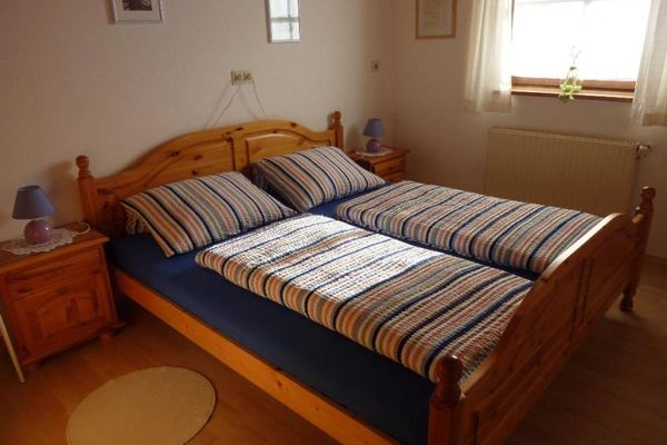 Kieferfarbenes Schlafzimmer mit Doppelbett, Nachtkästchen und Lampen. An der Wand hängen Bilder, am Fenster sind helle Vorhänge angebracht.